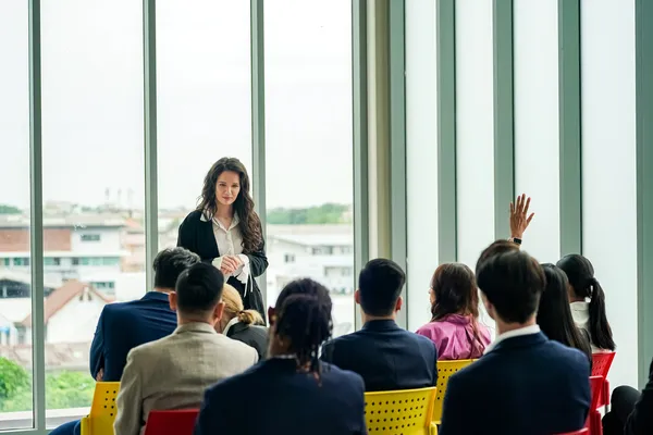 Ponente mujer frente a audiencia durante conferencias de coaching empresarial; una asistente levanta la mano para preguntar.