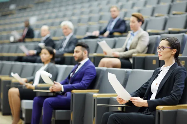 Profesionales atentos sentados en un auditorio, sosteniendo documentos en conferencias de coaching empresarial.